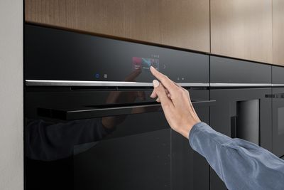 Une personne sélectionne les paramètres sur l'écran de commande tactile d'un four noir élégant, sur fond de cuisine moderne avec des armoires en bois.