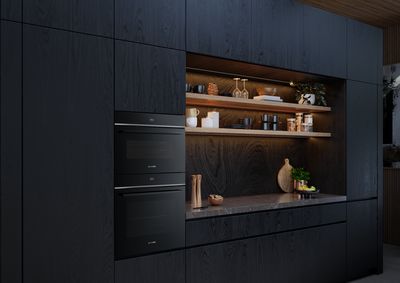 Cuisine moderne avec des armoires sombres, des fours encastrés, des étagères en bois pour exposer la vaisselle et la décoration, et un éclairage d'ambiance créant une atmosphère chaleureuse.
