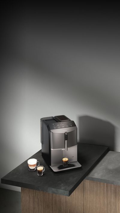 EQ300 stojí na príborníku s čiernou a sivou pracovnou doskou. Espreso stojí pod vylievacím otvorom, cappuccino je postavené vedľa neho.