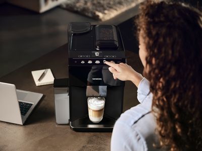 Woman is pressing the cappuccino button at the EQ500 coffee machine, a glass with coffee and milk foam already under the outlet.