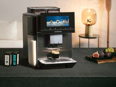 En EQ900 plus helautomatisk espressomaskin står på et svart bord, ved siden av den står et trefat med frukt, et kjøkkenhåndkle og til venstre ser man rengjøring- og avkalkingspatroner
