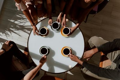 Widok z góry na okrągły stolik z białym blatem oraz ręce pięciu osób trzymających filiżanki z kawą.