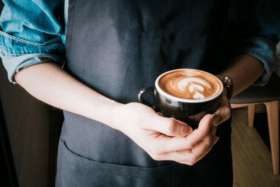 Barista w fartuchu trzyma obiema rękami kubek cappuccino