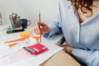 Kobieta w niebieskiej koszuli, trzymająca w dłoni ołówek, siedzi przy stole, na którym leżą paragony i faktury oraz różowy kalkulator.