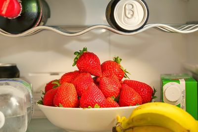 Biała miska pełna truskawek w lodówce, obok bananów i różnych napojów na półkach
