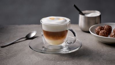 Cappuccino in einer Glastasse mit Milchschaum, Zucker und Gebäck.