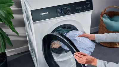 Hand opens Siemens washer door, revealing clothes inside; control panel visible above.