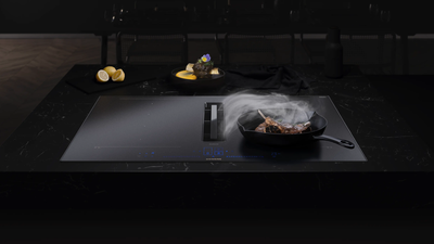 Een matte inductiekookplaat met geïntegreerde afzuiging bakt een steak in een gietijzeren pan terwijl inductionAir actief is en kookdampen afzuigt.