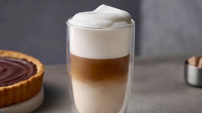 Ein isoliertes Glas mit Latte Macchiato, mit einem Kuchen und einem Zuckerschälchen im Hintergrund