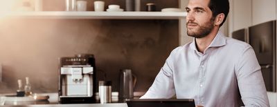 Junger Mann sitzt mit einer Tasse Kaffee in der Küche. Im Hintergrund ist ein EQ.6 Kaffeevollautomat