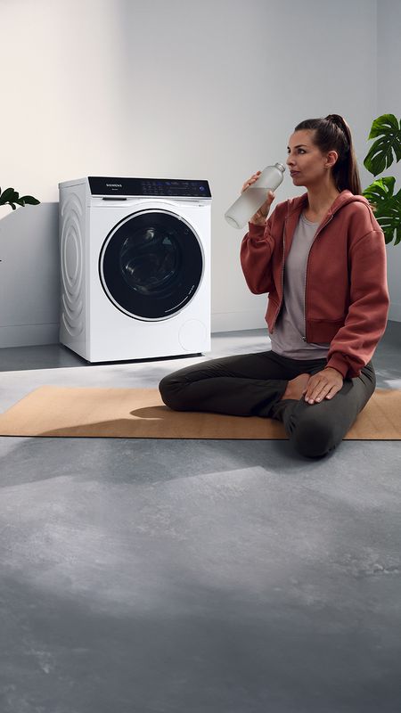 Ein Siemens Waschtrockner steht in einer Waschküche. Davor sitzt eine Frau im Schneidersitz und trinkt aus einer Wasserflasche.