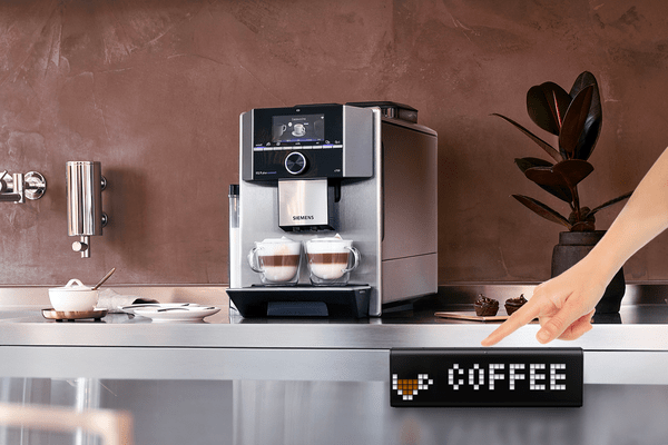 Zegar LaMetric, który wyświetla ikonkę kawy, znajduje się z przodu na blacie, w tle widać ekspres do kawy Siemens i dwie filiżanki cappuccino. 