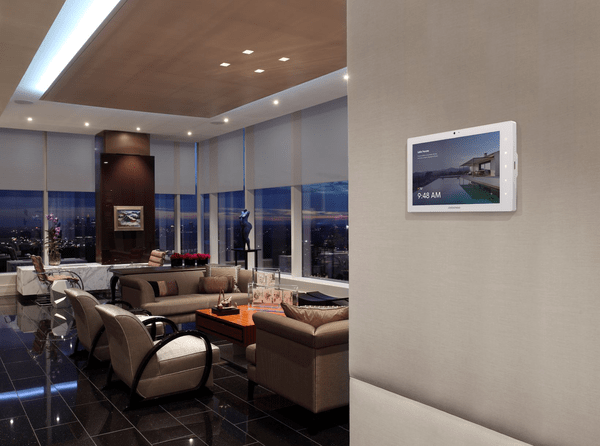 Crestron Smart Home panelis, piestiprināts pie sienas, fonā dzīvojamā istaba