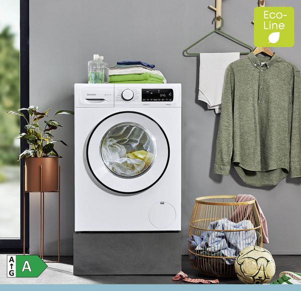 Eine Frontlader-Waschmaschine mit Wäsche steht neben einem Wäschekorb, einer Topfpflanze und einem hängenden Hemd in einem modernen Waschraum. Die Etiketten „Eco-Line“ und „Energieeffizienzklasse A“ sind sichtbar.