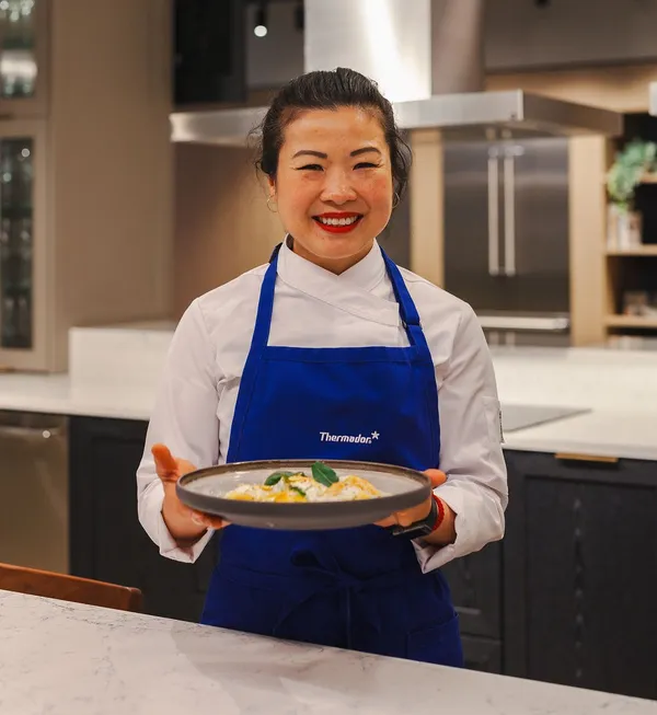 A chef in a white jacket and blue apron holds a plate of gourmet food, showcasing it against a modern kitchen backdrop