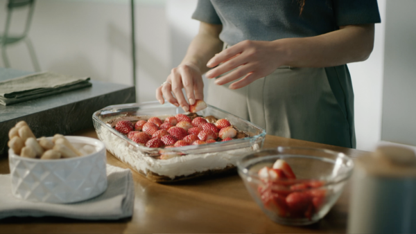 Una persona aggiunge fragole a un dessert in un piatto di vetro su un tavolo di legno, circondato da ciotole di frutta.