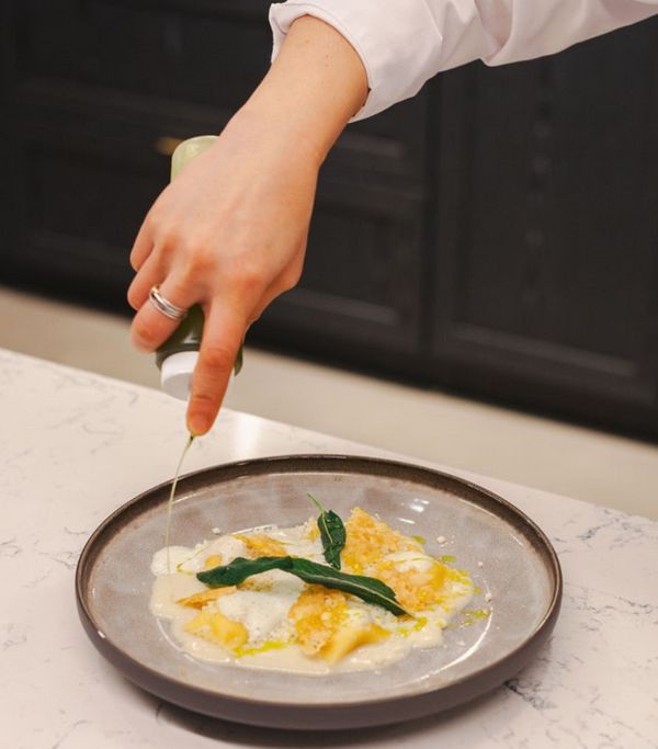 A chef's hand drizzles green sauce over a plate of pasta garnished with herbs and creamy sauce, on a marble countertop.