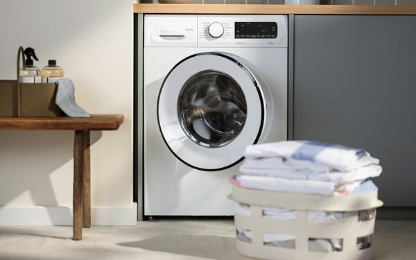 Ένα πλυντήριο ρούχων σε λευκό φωτισμένο χώρο με μπροστά του ένα καλάθι με φρεσκοπλυμένα σιδερωμένα ρούχα.