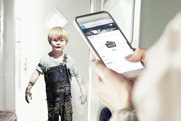 Vaikas purvinais drabužiais stovi prieškambaryje. Mama sustabdo skalbimo ciklą „Home Connect“ programėle.