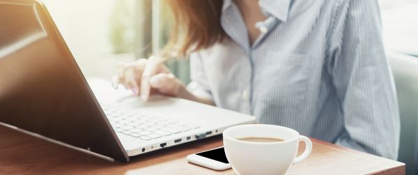 A young woman sitting at a laptop. The coffee next to her is freshly made by a Home Connect coffee machine. Thanks to an IFTTT recipe.