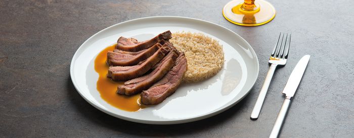 Un piatto di carne arrosto tagliata a fette con una ricca salsa, servita accanto a un riso soffice, accompagnata da forchetta e coltello su una superficie strutturata.