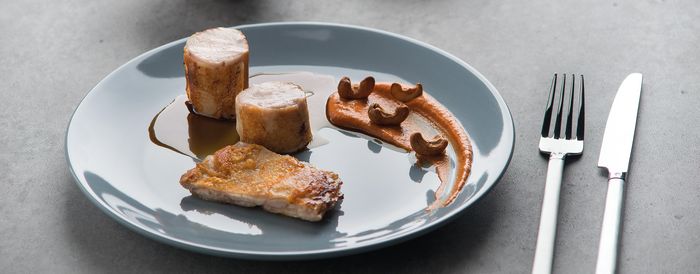 Un piatto gourmet al piatto con carne di maiale scottata, tagli di carne cilindrici, una salsa morbida e anacardi sparsi, accompagnato da eleganti posate.