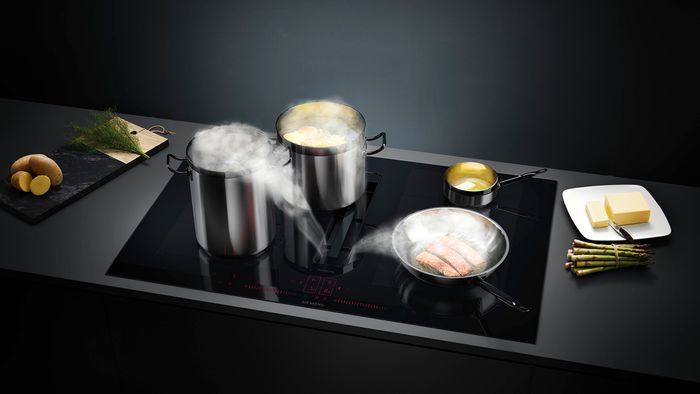 Food cooking in numerous pots and pans on venting hob with steam going down into it