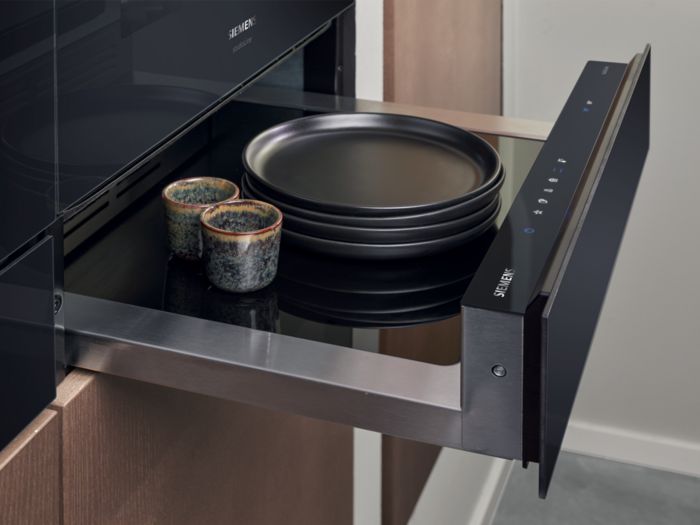 Un elegante cassetto da cucina Siemens contiene due tazze decorative e piatti neri, con un moderno pannello a sfioramento per il controllo.