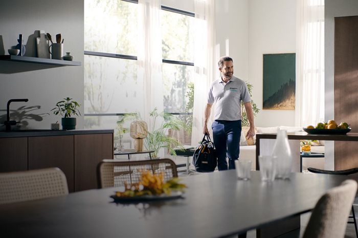 Mies astuu moderniin keittiöön mustaa laukkua kantaen, kasvien ja kirkkaiden kasvien ympäröimänä.