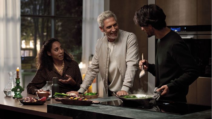 Zwei Männer und eine Frau stehen in einer offenen Küche um eine Kochinsel mit einem Siemens Kochfeld mit Dunstabzug.