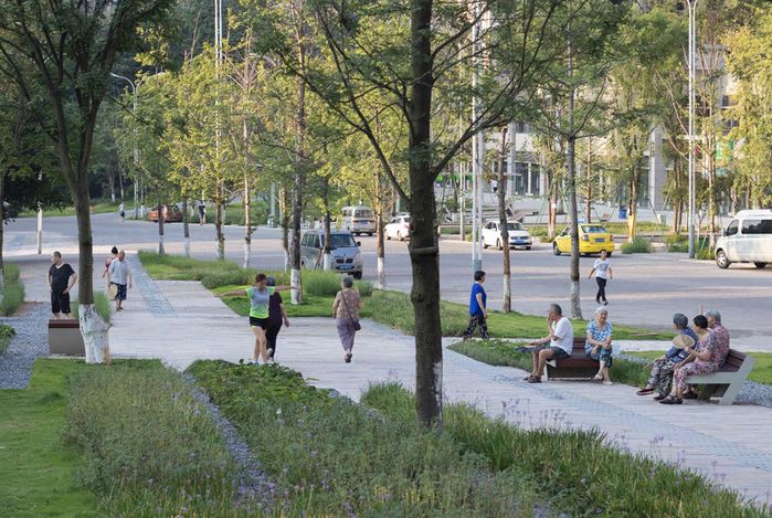 Urban scene with people walking and sitting in a park-like setting surrounded by greenery.