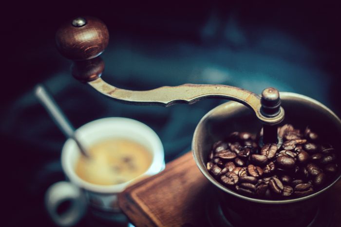 Ręczny młynek do kawy z kolbą i ziarnami kawy, w tle rozmazana filiżanka kawy z pianką. 