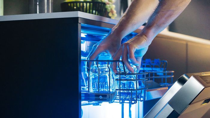 Manos cargando lavavajillas con cristalería limpia, iluminación interior azul.