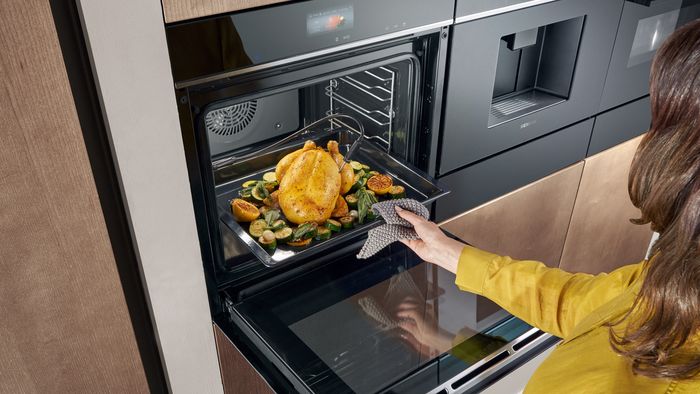Eine Frau schiebt ein Backblech mit Hühnchen und Gemüse in den Siemens Backofen.