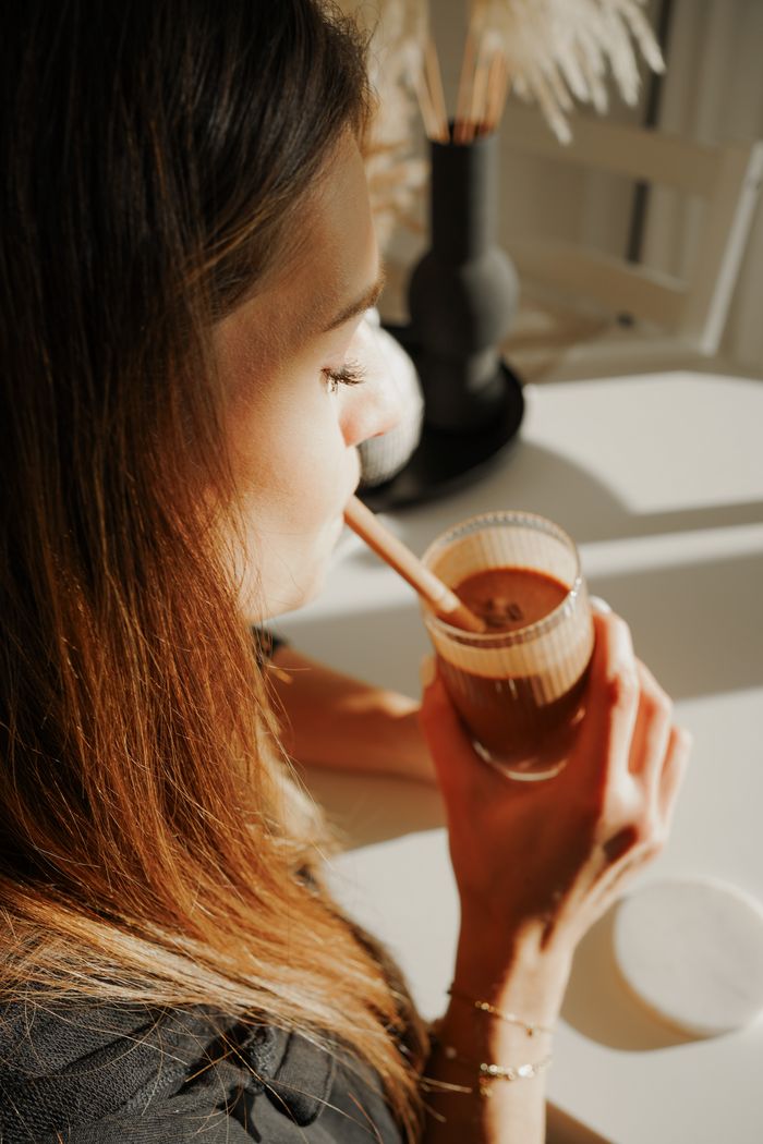 Het midden van de afbeelding: Een klein bordje met koekjes en daarnaast een glas met ijskoffie met halfbevroren schuim. Wazig op de achtergrond is Heike bezig met de volgende koffie.