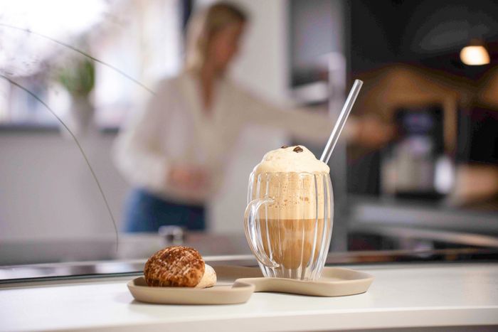 Het midden van de afbeelding: Een klein bordje met koekjes en daarnaast een glas met ijskoffie met halfbevroren schuim. Wazig op de achtergrond is Heike bezig met de volgende koffie.