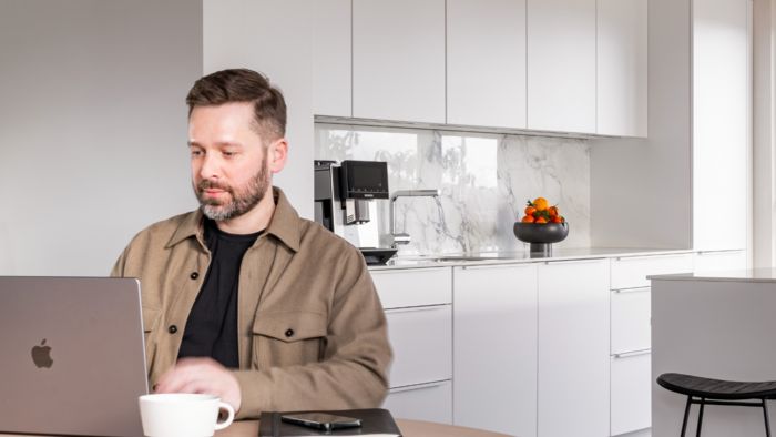 Dominik est assis à la table à manger et travaille sur son ordinateur. Sa cuisine est à l'arrière-plan. Une tasse de café est posée sur la table à côté de lui. Boire du café en travaillant fait partie de ses habitudes.