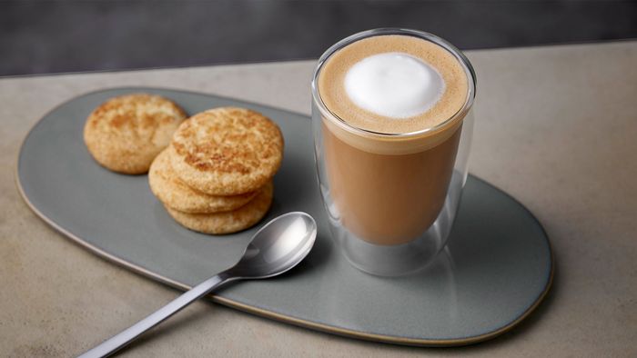 Eine Tasse mit Flat White Kaffee auf einem Teller mit Gebäck und Löffel.