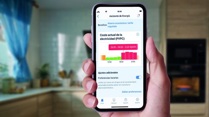 Una persona sostiene un smartphone en el que se muestra una aplicación de asistente energético que muestra en tiempo real los costes de la electricidad y consejos para ahorrar en una cocina.