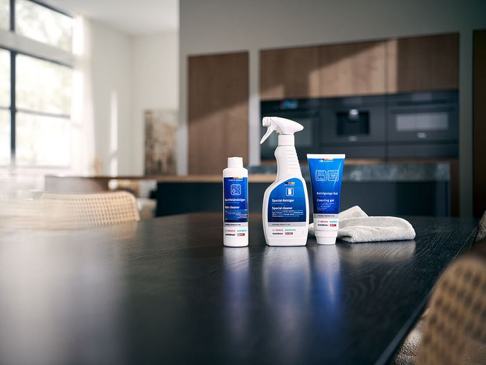 Tre prodotti per la pulizia con etichette blu, tra cui un flacone spray e due tubetti, sono disposti su un tavolo da cucina in legno scuro con un panno.