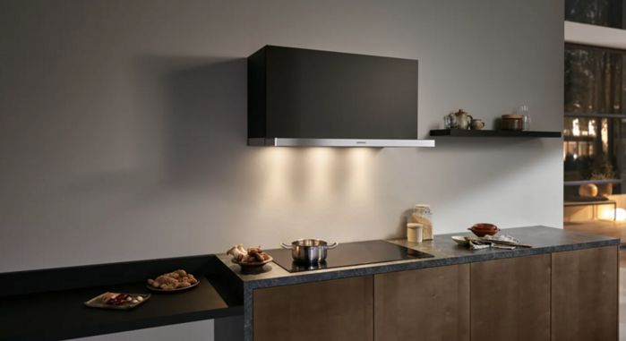 Cocina moderna con una elegante campana extractora integrada Siemens en negro y una placa de inducción con una olla, rodeada de encimera con ingredientes y platos.