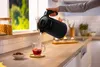 Person gießt heißes Wasser aus einem Wasserkocher in eine Glasteetasse auf einem Holztisch.
