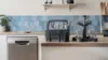 Modern kitchen countertop with a dishwasher, cutlery holder, wine glass rack, blue tiled backsplash, and minimal decor.