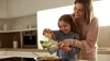Mother and daughter smiling while grating cheese over a dish in a modern kitchen.