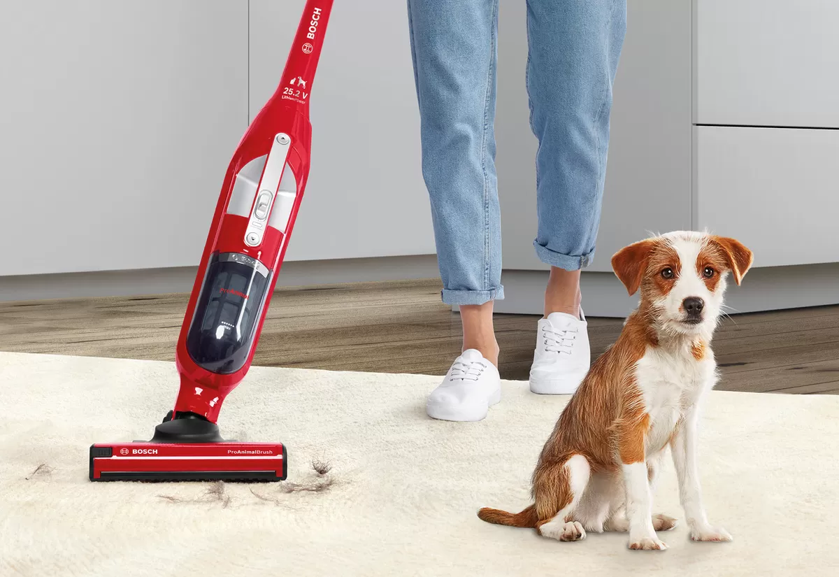 Se muestra una aspiradora Bosch roja sobre una alfombra, diseñada para el pelo de mascotas, cerca de un perro pequeño sentado, con una cocina moderna de fondo.