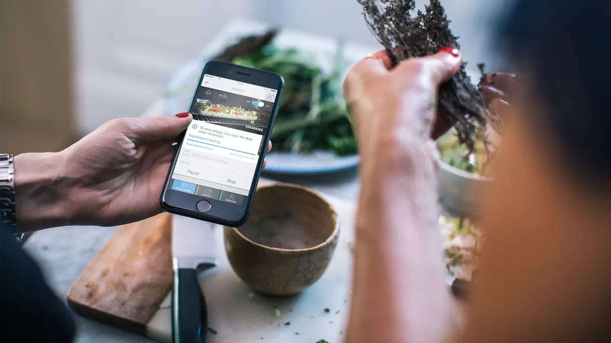 Två kvinnohänder syns över en köksbänk där mat förbereds och en iPhone med Home Connect-appen är öppen. Appen visar recept och tips.