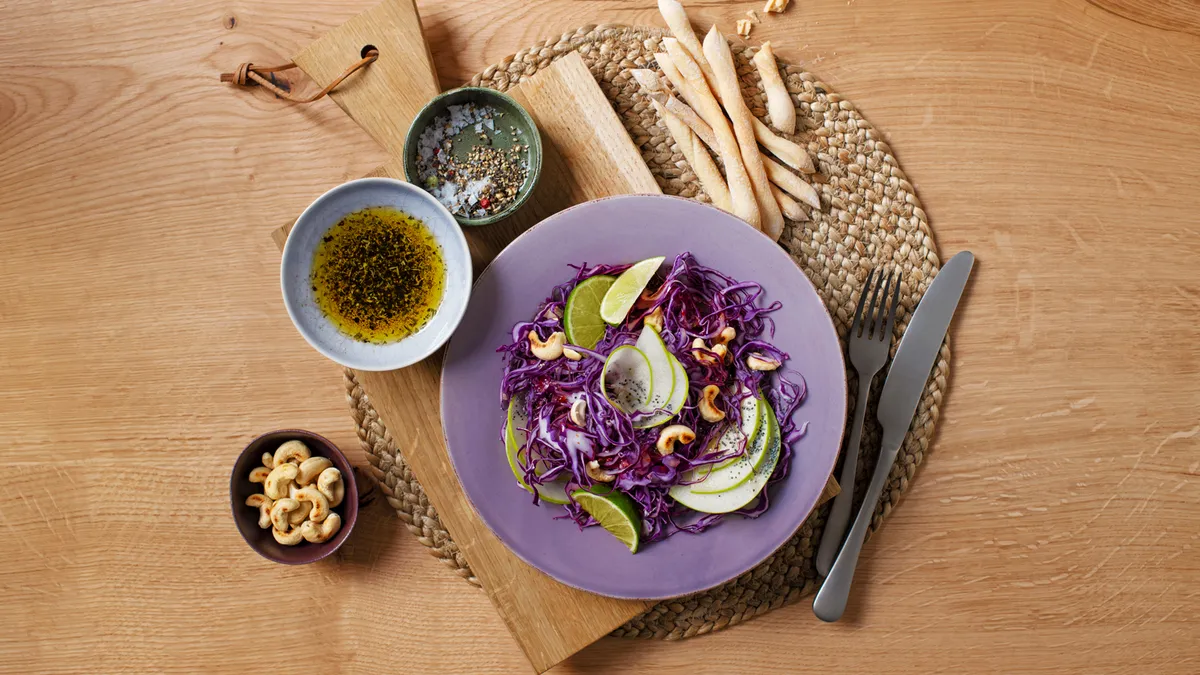 منظر من أعلى لملفوف أحمر وسلطة تفاح مع الكاجو وشرائح الليمون وبذور الخشخاش على طبق أرجواني، يقدم مع صلصة جانبية والكاجو وأعواد الخبز على سطح خشبي.