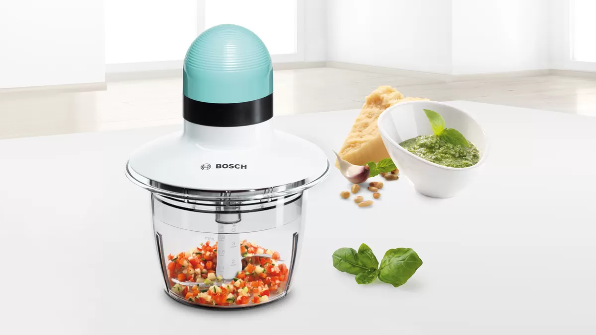 مفرمة طعام كهربائية من شركة بوش تحتوي على خضراوات مقطعة في وعاء خاص بها، بجانب وعاء من البيستو، وجبن البارميزان، وأوراق الريحان، والصنوبر، وفصوص الثوم على سطح أبيض.