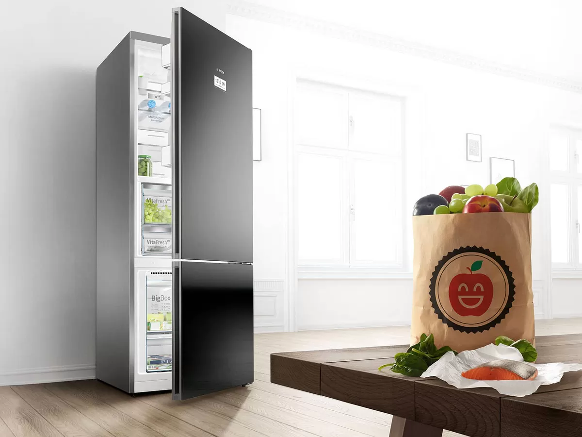 Moderno frigorífico negro con las puertas abiertas dejando ver productos frescos, junto a una bolsa de la compra llena de frutas y verduras sobre una mesa de madera.