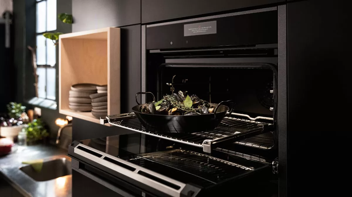 Ένας άνδρας τοποθετεί ένα ταψί με πράσινα λαχανικά σε ανοικτό μόνο φούρνο σε μια μαύρη κουζίνα